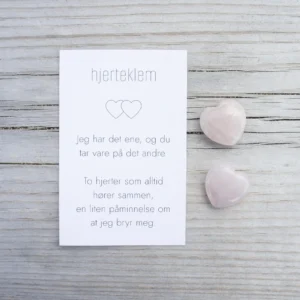 Et kort med teksten “hjertklem” og to rosa steinhjerter ved siden av, på lyst treverk.