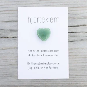 Kort med teksten “hjerteklem” og en grønn hjerteformet stein limt på, lagt på lys trebakgrunn. Kortet sier: “Her er en hjerteklem som du kan ha i lommen din. En liten påminnelse om at jeg alltid er her for deg.”