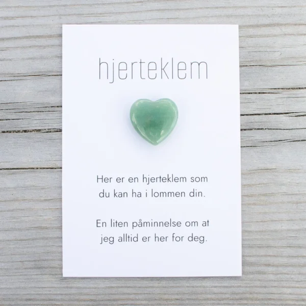 Kort med teksten “hjerteklem” og en grønn hjerteformet stein limt på, lagt på lys trebakgrunn. Kortet sier: “Her er en hjerteklem som du kan ha i lommen din. En liten påminnelse om at jeg alltid er her for deg.”