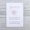 Kort med teksten “hjerteklem” og en rosa hjerteformet stein limt på, lagt på lys trebakgrunn. Kortet sier: “Her er en hjerteklem som du kan ha i lommen din. En liten påminnelse om at jeg alltid er her for deg.”