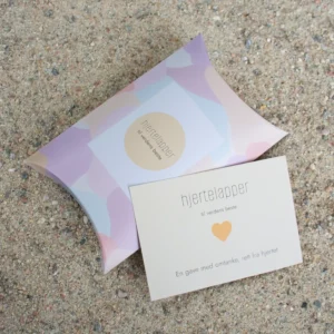 Hjertelapp med oransje hjerte og teksten “til verdens beste”, fotografert sammen med matchende gaveeske på sand.