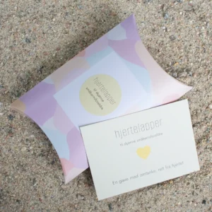 Hjertelapp med gult hjerte og teksten “til skjønne småbarnsforeldre”, fotografert sammen med matchende gaveeske på sand.