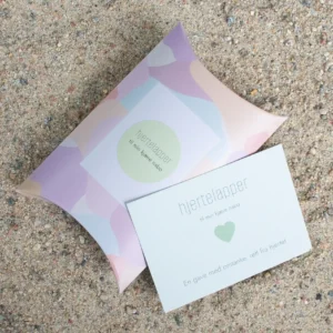 Hjertelapp med grønt hjerte og teksten “til min kjære nabo”, fotografert sammen med matchende gaveeske på sand.