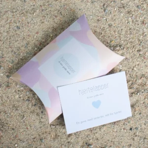 Hjertelapp med blått hjerte og teksten “til min gode venn”, fotografert sammen med matchende gaveeske på sand.