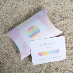 Hjertelapp med regnbuehjerter og teksten “fra meg til deg”, sammen med en matchende gaveeske med holografisk klistremerke, plassert på sand.