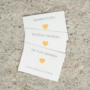 Tre lys oransje hjertelapper med teksten “Mimrestund”, “Koselig middag” og “En tur sammen”, liggende på sand. Hver lapp har et oransje hjerte og enkel, sort tekst.