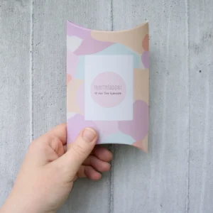 Gaveeske med pastellfarget mønster og teksten “Hjertelapper – til min fine kjæreste”, holdt i hånden foran en lys betongvegg.