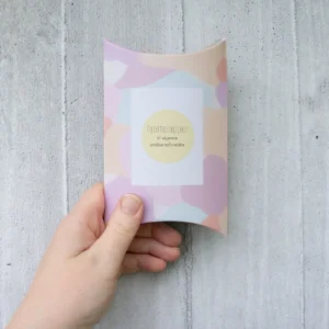 Gaveeske med pastellfarget mønster og teksten “Hjertelapper – til skjønne småbarnsforeldre”, holdt i hånden foran en lys betongvegg.