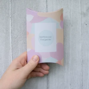 Gaveeske med pastellfarget mønster og teksten “Hjertelapper – til min gode venn”, holdt i hånden foran en lys betongvegg.