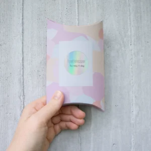 Gaveeske med pastellfarget mønster og teksten “Hjertelapper – fra meg til deg”, holdt i hånden foran en lys betongvegg.