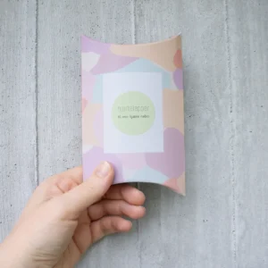 Gaveeske med pastellfarget mønster og teksten “Hjertelapper – til min kjære nabo”, holdt i hånden foran en lys betongvegg.