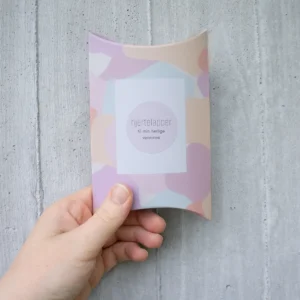 Gaveeske med pastellfarget mønster og teksten “Hjertelapper – til min herlige venninne”, holdt i hånden foran en lys betongvegg.