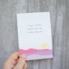 Kort med teksten «Du gjør verden bedre bare ved å være deg selv», og illustrasjon av rosa fjell og sol. Holdt i en hånd foran en lys betongvegg.