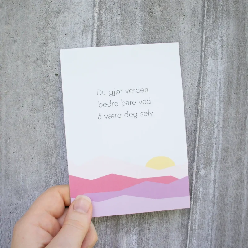 Kort med teksten «Du gjør verden bedre bare ved å være deg selv», og illustrasjon av rosa fjell og sol. Holdt i en hånd foran en lys betongvegg.