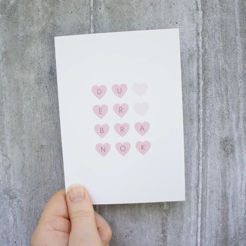 Kort med rosa hjerter som danner setningen «DU ER BRA NOK», holdt i en hånd foran en lys betongvegg.