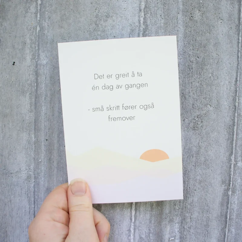 Kort med teksten «Det er greit å ta én dag av gangen – små skritt fører også fremover» og en enkel soloppgang-illustrasjon nederst. Holdt i en hånd foran lys betongvegg.