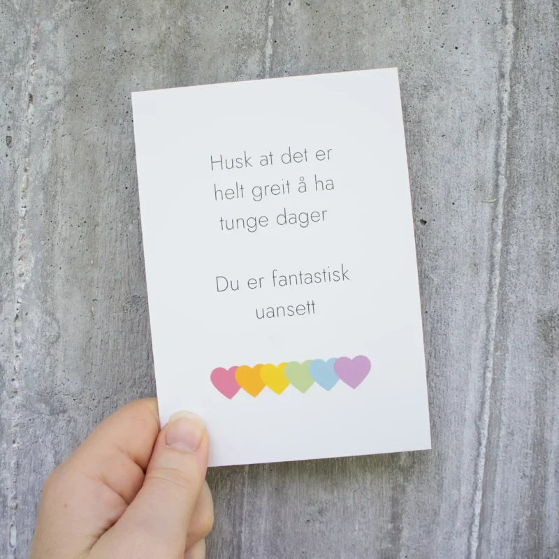 Kort med teksten “Husk at det er helt greit å ha tunge dager – du er fantastisk uansett”, holdt foran en lys betongvegg. Nederst på kortet er seks hjerter i regnbuefarger.
