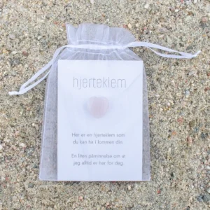 En hvit gavepose med kort og ett rosa krystallhjerte foran, plassert på sand.