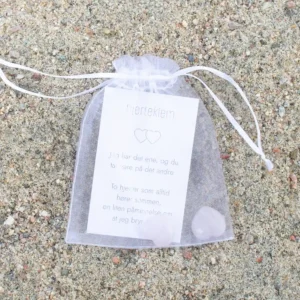 En hvit gavepose med kort og to rosa hjerter i krystall, fotografert på sand.