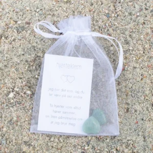 En hvit gavepose med kort og to grønne hjerter i krystall, plassert på sand.