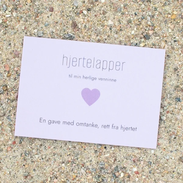 Kort med lilla hjerte og teksten «hjertelapper – til min herlige venninne», fotografert på sand.
