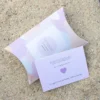 Hjertelapp med lilla hjerte og teksten “til min herlige venninne”, fotografert sammen med matchende gaveeske på sand.