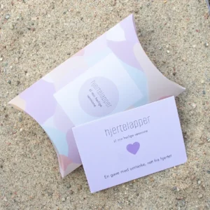 Hjertelapp med lilla hjerte og teksten “til min herlige venninne”, fotografert sammen med matchende gaveeske på sand.