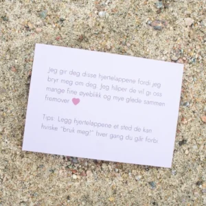 Kort med tekst om hjertelapper og et rosa hjerte, liggende på sand. Teksten uttrykker omtanke og et tips om hvordan lappene kan brukes i hverdagen.
