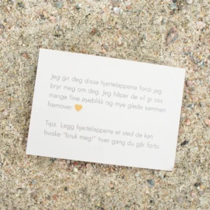 Kort med tekst om hjertelapper og et oransje hjerte, liggende på sand. Teksten uttrykker omtanke og et tips om hvordan lappene kan brukes i hverdagen.