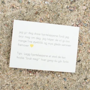 Kort med tekst om hjertelapper og et gult hjerte, liggende på sand. Teksten uttrykker omtanke og et tips om hvordan lappene kan brukes i hverdagen.