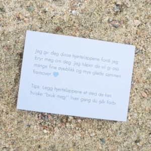 Kort med tekst om hjertelapper og et blått hjerte, liggende på sand. Teksten uttrykker omtanke og et tips om hvordan lappene kan brukes i hverdagen.