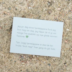 Kort med tekst om hjertelapper og et grønt hjerte, liggende på sand. Teksten uttrykker omtanke og et tips om hvordan lappene kan brukes i hverdagen.