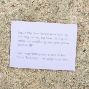 Kort med tekst om hjertelapper og et lilla hjerte, liggende på sand. Teksten uttrykker omtanke og et tips om hvordan lappene kan brukes i hverdagen.