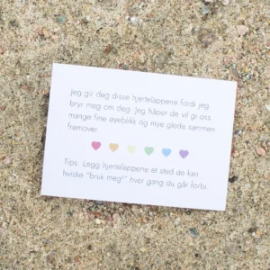 Kort med tekst om hjertelapper og seks fargede hjerter, liggende på sand. Teksten uttrykker omsorg og oppmuntrer til bruk av hjertelappene i hverdagen.