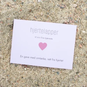 Kort med rosa hjerte og teksten «hjertelapper – til min fine kjæreste», fotografert på sand.