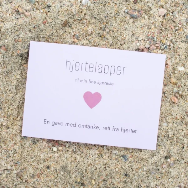 Kort med rosa hjerte og teksten «hjertelapper – til min fine kjæreste», fotografert på sand.