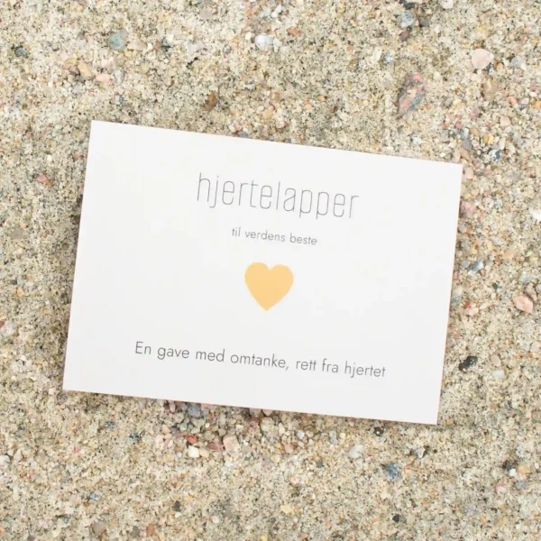 Kort med oransje hjerte og teksten «hjertelapper – til verdens beste», fotografert på sand.