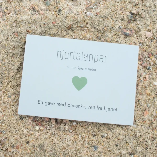 Kort med grønt hjerte og teksten «hjertelapper – til min kjære nabo», fotografert på sand.