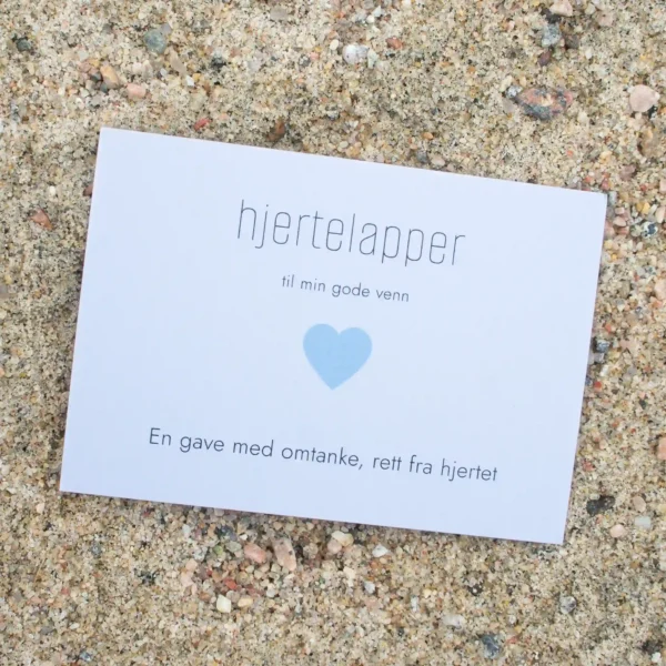 Kort med blått hjerte og teksten «hjertelapper – til min gode venn», fotografert på sand.