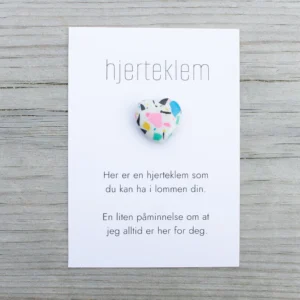 Kort med teksten “hjerteklem” og en fargerik hjertestein limt på, lagt på lys trebakgrunn. Kortet sier: “Her er en hjerteklem som du kan ha i lommen din. En liten påminnelse om at jeg alltid er her for deg.”