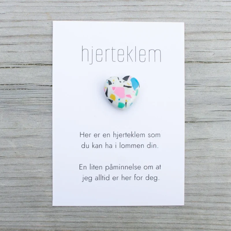 Kort med teksten “hjerteklem” og en fargerik hjertestein limt på, lagt på lys trebakgrunn. Kortet sier: “Her er en hjerteklem som du kan ha i lommen din. En liten påminnelse om at jeg alltid er her for deg.”