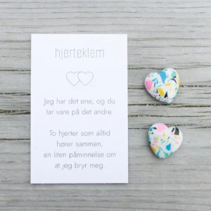 Et kort med teksten “hjertklem” og to fargerike steinhjerter ved siden av, på lyst treverk.