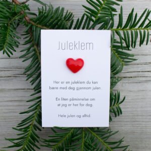 Kortet “Juleklem” med rødt hjerte, omkranset av grønne grener på lyst underlag.