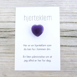 Kort med teksten “hjerteklem” og en dyp lilla hjertestein limt på, lagt på lys trebakgrunn. Kortet sier: “Her er en hjerteklem som du kan ha i lommen din. En liten påminnelse om at jeg alltid er her for deg.”
