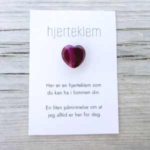Kort med teksten “hjerteklem” og en dyp rosa hjertestein limt på, lagt på lys trebakgrunn. Kortet sier: “Her er en hjerteklem som du kan ha i lommen din. En liten påminnelse om at jeg alltid er her for deg.”