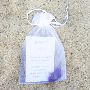En hvit gavepose med kort og to krystallhjerter i dyp lilla, fotografert på sand.