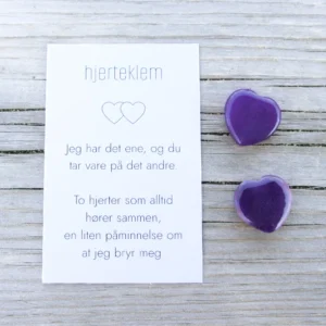 Et kort med teksten “hjertklem” og to dyp lilla steinhjerter ved siden av, på lyst treverk.