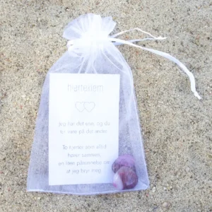 En hvit gavepose med kort og to krystallhjerter i dyp rosa, fotografert på sand.