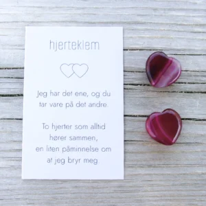 Et kort med teksten “hjertklem” og to dyp rosa steinhjerter ved siden av, på lyst treverk.